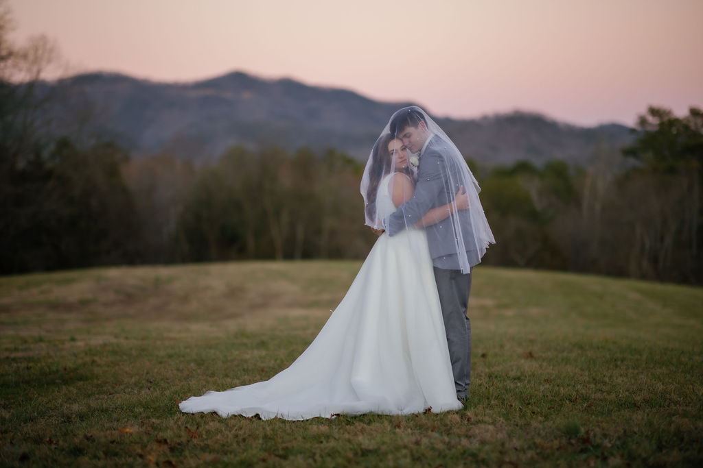 bride adn groom holding eachother in a field right after the sunsets while under the brides veil for their Gatlinburg elopement