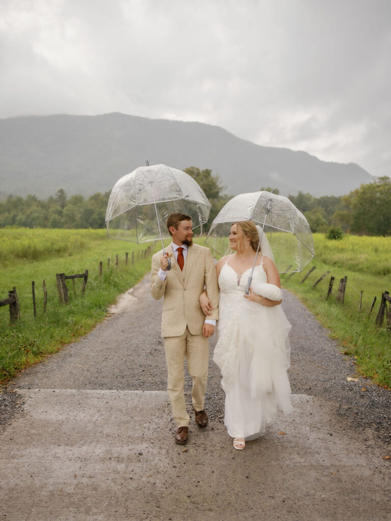 rainy wedding day in the mountains with bride and groom holding clear umbrellas while walking on a path in cades cove