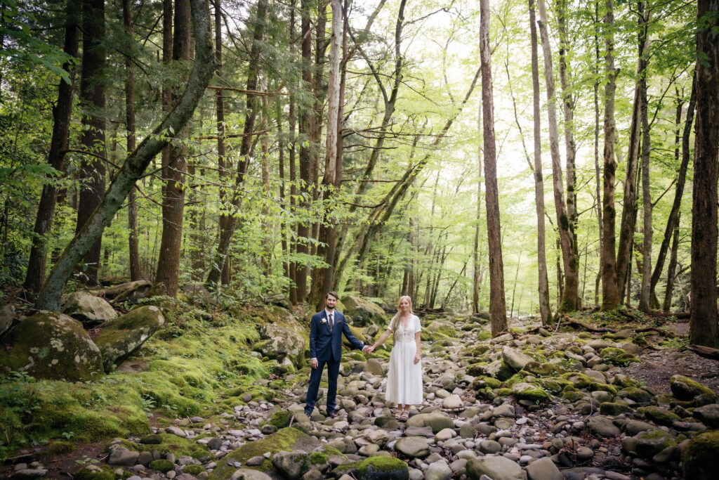 bride and groom holding hands and standing in a river bed full of rocks in the woods near Spence Cabin