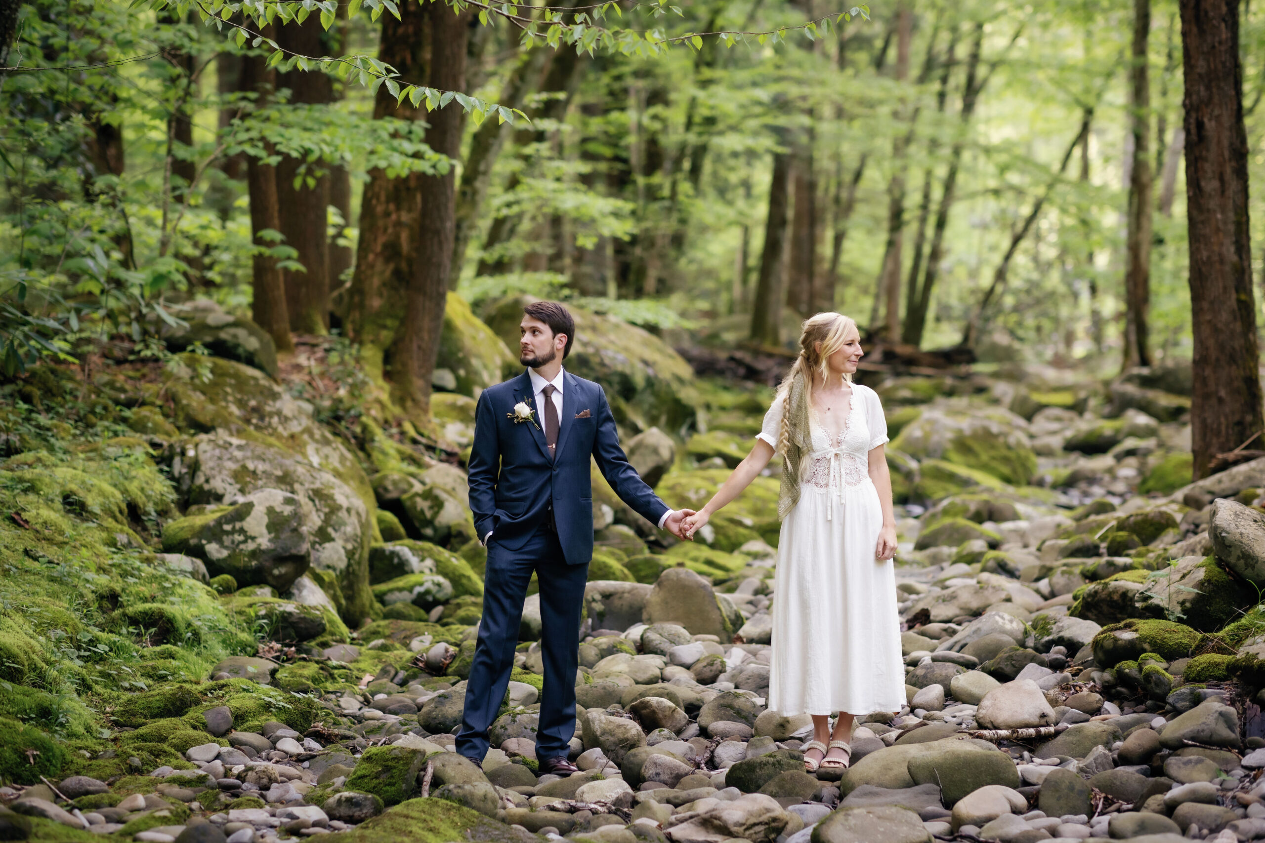 bride and groom hold hands and look opposite directions as they stand on river rocks in the woods of the Smokies