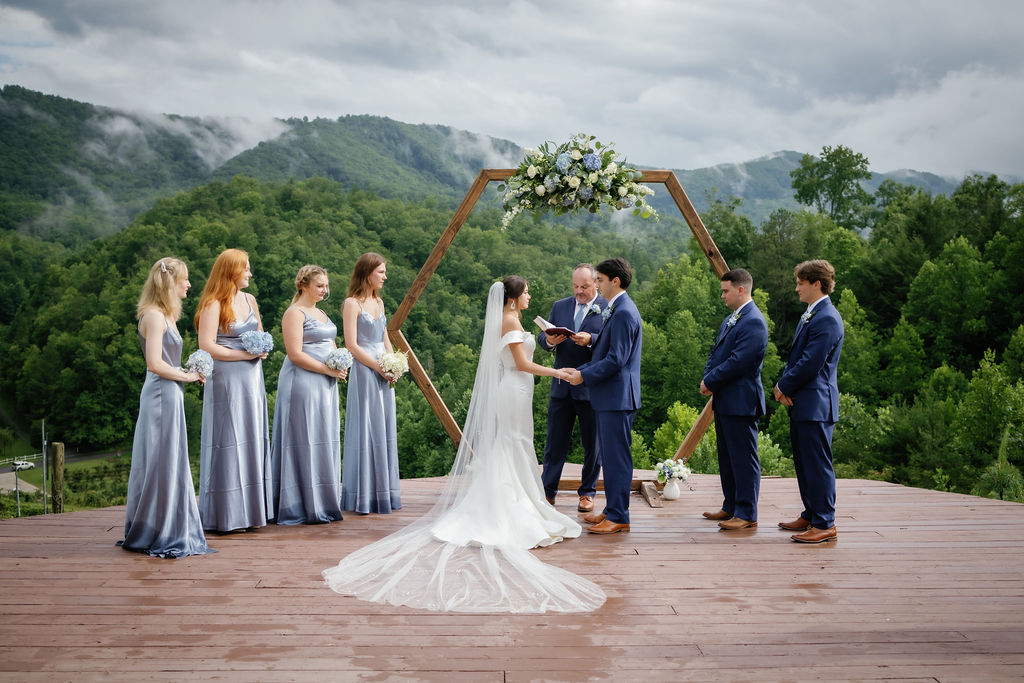 outdoor wedding ceremony for a Smoky Mountain elopement with bride and groom holding hands on a deck surrounded by their bridal party while saying their vows