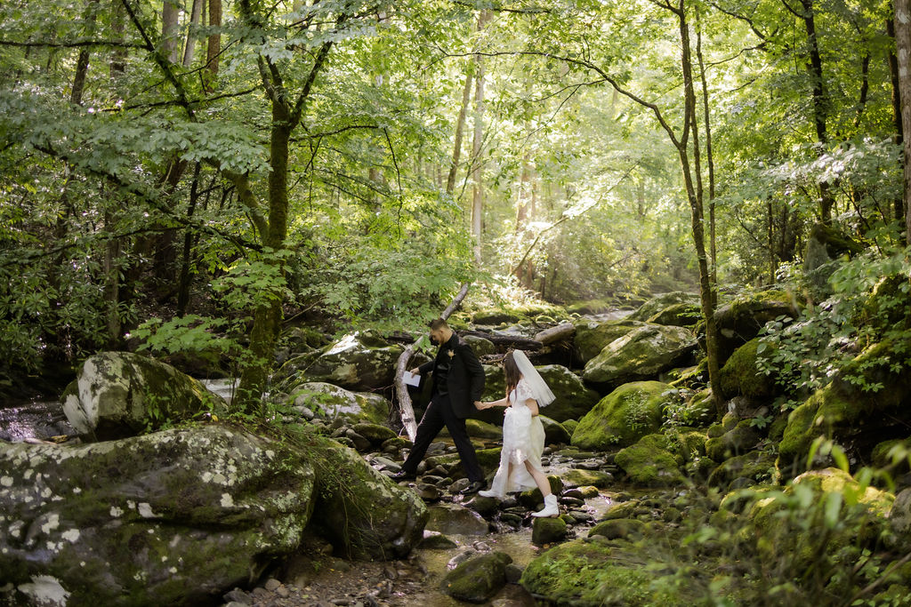 photographers in gatlinburg tn captures candid wedding photo of bride and groom holding hands as they climb over rocks together in the woods near Ely's Mill