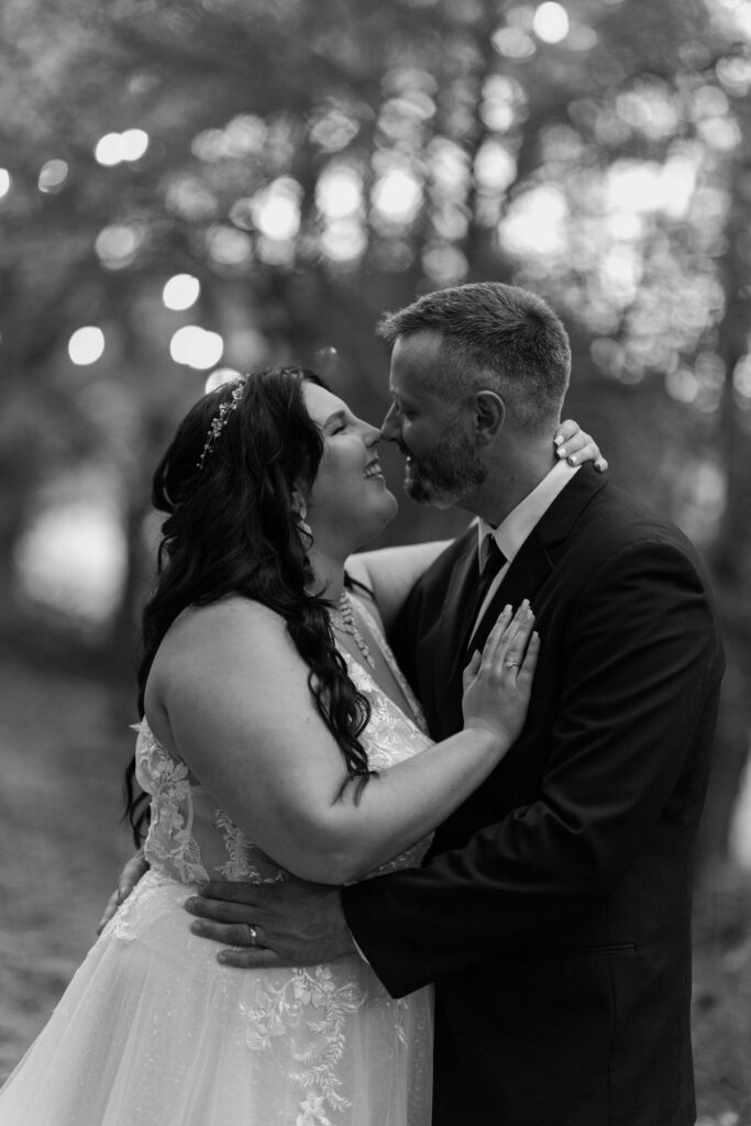 A black-and-white close-up captures a joyful bride and groom in a tender embrace, their noses nearly touching as they smile at each other. The bride's hand rests gently on the groom’s chest, showcasing her engagement ring, while the groom holds her close, both radiating happiness and intimacy. Soft lights twinkle in the blurred background, adding a dreamy ambiance that highlights the romance of eloping to Gatlinburg, ideal for couples seeking photographers in Gatlinburg, TN.