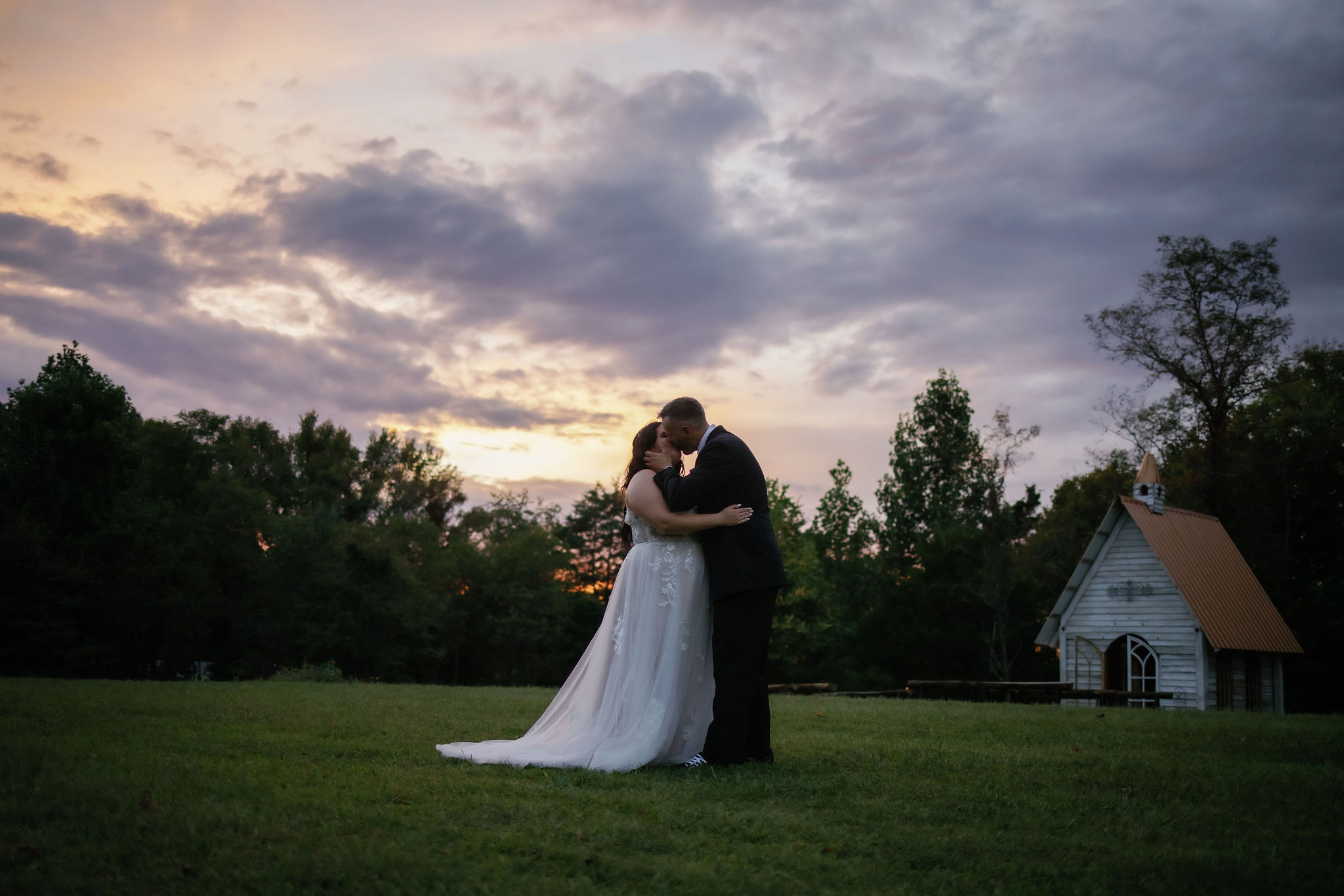 photographers in gatlinburg tn captures Bride and groom sharing a kiss on a grassy field at sunset, with a small chapel in the background. The bride wears a flowing lace wedding gown, and the groom is dressed in a dark suit. The sky is filled with soft clouds, creating a romantic, peaceful atmosphere in the scenic outdoor setting