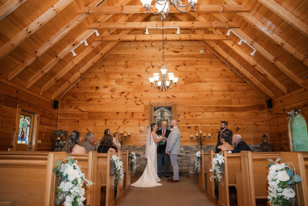 The couple exchanges vows during a touching Chapel in the Glen elopement in Gatlinburg, Tennessee. The warm, pine wood interior of the chapel provides a cozy and intimate atmosphere, with vaulted ceilings and exposed beams creating a rustic elegance. Guests are seated on wooden pews adorned with white floral and greenery arrangements, perfectly complementing the chapel’s natural charm.