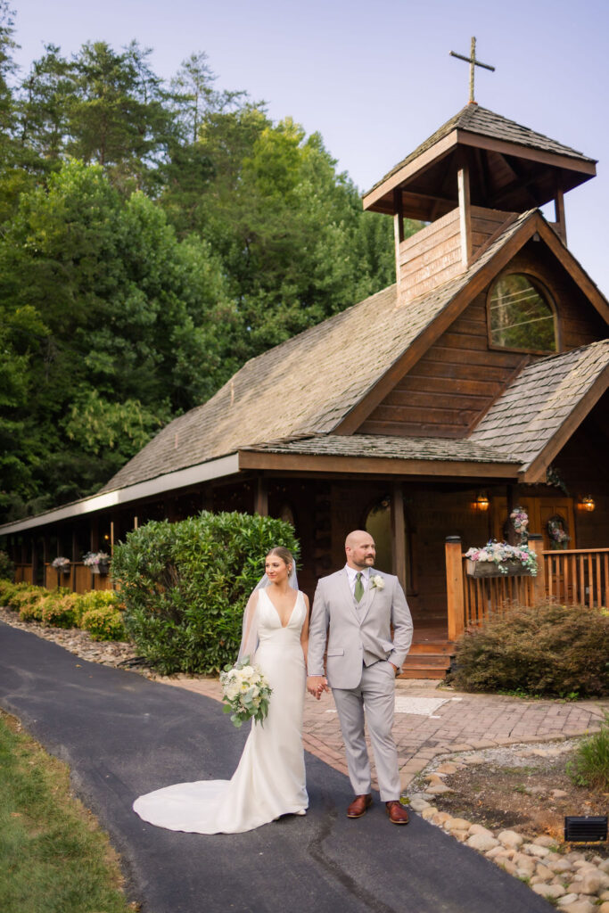 The bride and groom walk hand in hand along a smooth blacktop path in front of the charming Chapel in the Glen, a perfect venue for a Gatlinburg elopement. The rustic wooden chapel stands proudly in the background, with its steep roofline, cross-topped steeple, and flower-adorned railings. Vibrant greenery, including neatly trimmed bushes and colorful flower beds, lines the walkway, enhancing the picturesque scene. The bride’s flowing white gown and bouquet of fresh blooms elegantly contrast with the natural tones of the wood and foliage, creating a timeless and romantic moment.