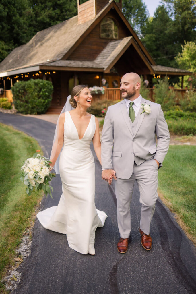 A joyful bride and groom hold hands as they walk along a curved, paved pathway in front of the charming Chapel in the Glen. The bride glows in a sleek white gown with a deep V neckline and a flowing veil, holding a lush bouquet of white roses and greenery. The groom looks dashing in a light gray suit accented with a green tie and boutonniere. Behind them, the wooden chapel features a steeply pitched roof, twinkling string lights, and vibrant greenery, blending seamlessly with the tranquil surroundings of this Gatlinburg elopement venue.