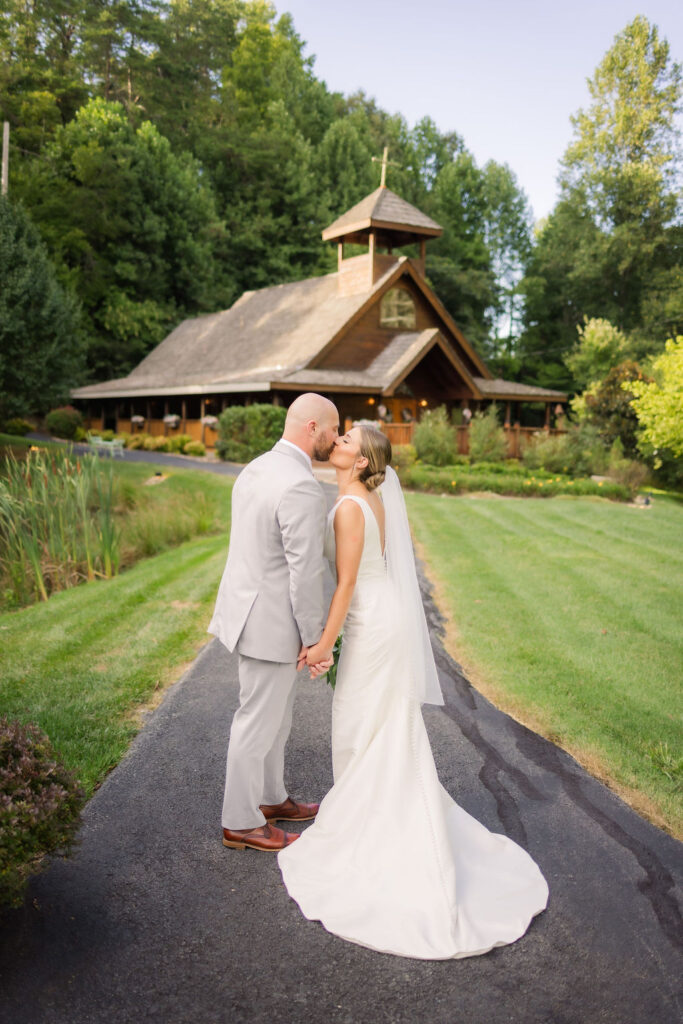 A couple shares a romantic kiss on a paved pathway leading to the picturesque Chapel in the Glen during their Gatlinburg elopement. Surrounded by lush greenery, the chapel’s rustic wooden structure with its charming steeple and wrap-around porch creates a perfect backdrop. To the left, a pond bordered by reeds and a neatly manicured lawn adds to the serene setting. The bride’s elegant white gown and veil softly contrast with the natural tones of the landscape, enhancing the magical atmosphere of the moment.