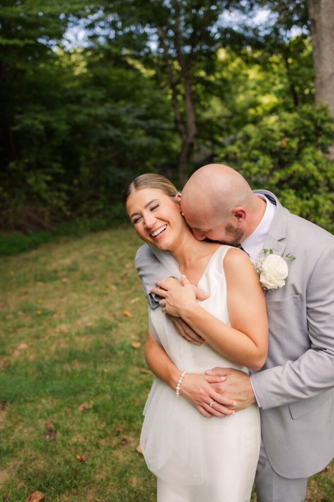 he groom playfully embraces his bride in a candid moment of laughter and joy during their Gatlinburg elopement. Set against a backdrop of lush greenery and mature trees, this lighthearted capture perfectly highlights the couple’s love and connection. The bride’s radiant smile, her pearl bracelet, and simple yet elegant gown complement the natural surroundings, while the groom’s tender expression and rose boutonniere add a romantic touch.