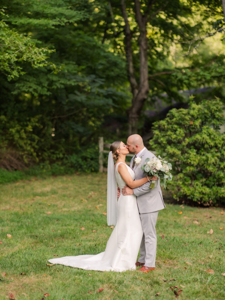 The bride and groom share a tender kiss during their Chapel in the Glen elopement, standing in a serene outdoor setting surrounded by lush greenery. The bride's sleek satin gown and flowing veil create a timeless elegance, complemented by the soft white and green tones of her bouquet. The groom’s light gray suit and warm brown shoes harmonize with the natural tones of the grassy lawn and vibrant foliage.