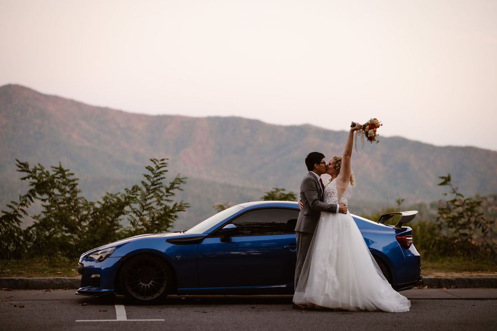 Gatlinburg photographer captures bride and groom kissing in front of a sports car with the mountains in the distance as the bride holds her bouquet in the air