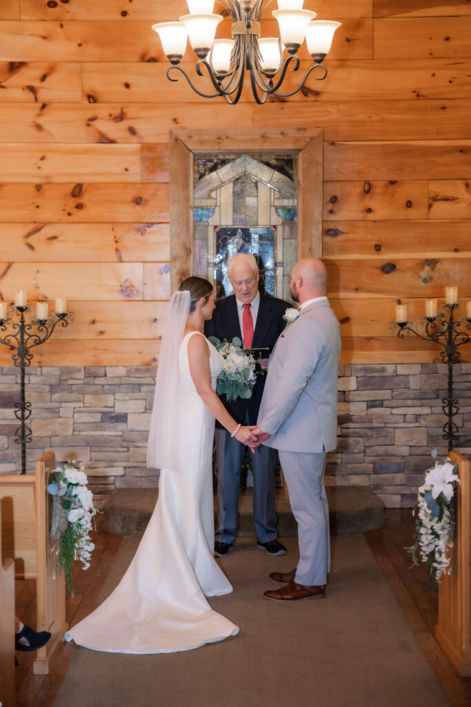 A heartfelt moment during a Chapel in the Glen elopement ceremony in Gatlinburg, Tennessee, as the couple stands hand in hand before the officiant. The chapel’s warm wooden walls and a central stained-glass window create an intimate, rustic ambiance. On either side of the couple, black wrought-iron candelabras adorned with glowing pillar candles add a romantic touch. The pews are decorated with white florals and greenery, enhancing the cozy elegance of this special setting