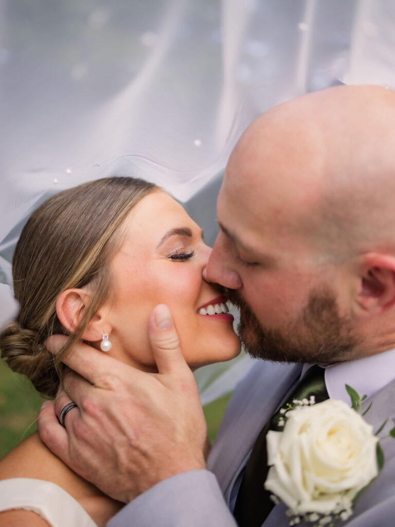 A tender close-up of a bride and groom sharing a kiss, framed delicately by the soft drape of the bride's pearl-studded veil. The bride’s hair is swept into an elegant low bun, showcasing her radiant smile and classic pearl earrings. The groom’s hand gently cradles her face, highlighting his wedding band and the couple’s deep connection. A white rose boutonniere on the groom’s light gray suit adds a timeless touch, completing this intimate moment from a romantic Gatlinburg elopement.