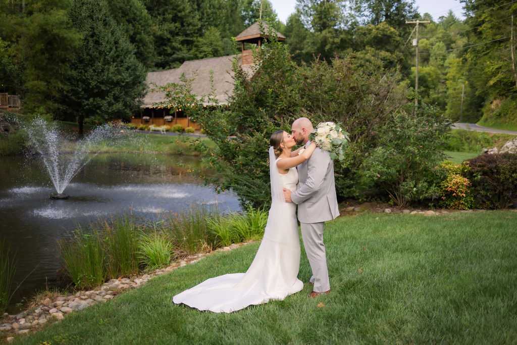 A newlywed couple shares a romantic embrace on a lush green lawn beside a serene pond with a fountain spraying water gracefully into the air. The bride’s fitted, elegant white gown cascades onto the grass, while she holds onto her groom, who wears a light gray suit and clasps a bouquet of white flowers. Surrounding the scene, vibrant shrubbery and flowering plants add bursts of color, with the rustic wooden Chapel in the Glen visible in the background, nestled among tall trees.