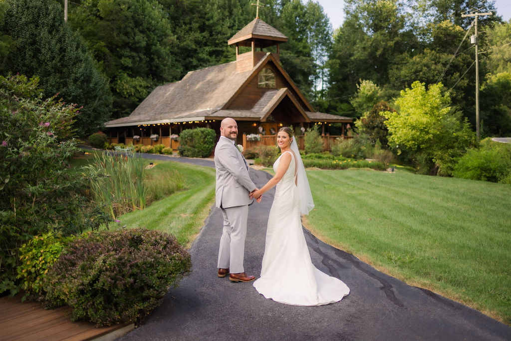 A newlywed couple holds hands and smiles back at the camera, standing on the path leading to the Chapel in the Glen, embodying the intimate charm of a Chapel in the Glen elopement amidst Gatlinburg’s natural beauty