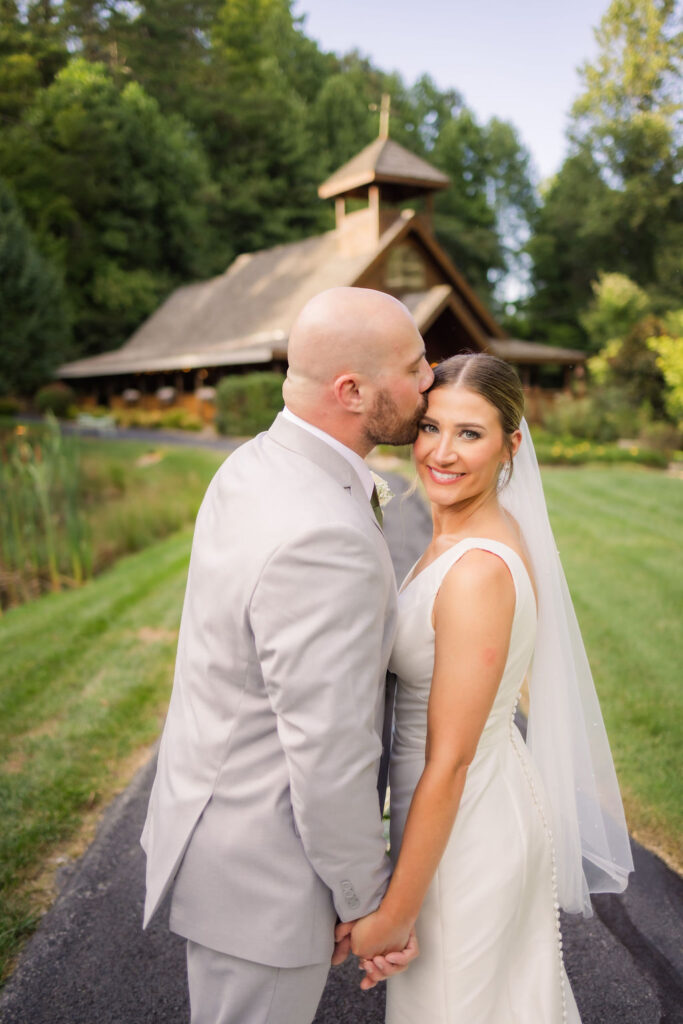 A groom in a sharp light gray suit gently kisses his bride's temple, creating a tender moment as she smiles radiantly at the camera. The bride, in a sleek, button-back white gown and delicate veil, holds hands with her partner, exuding elegance. Behind them, a paved pathway winds through lush green grass, leading to the picturesque wooden Chapel in the Glen, nestled among dense, towering trees. The tranquil surroundings and thoughtful landscaping add to the romantic ambiance of this Gatlinburg elopement setting.