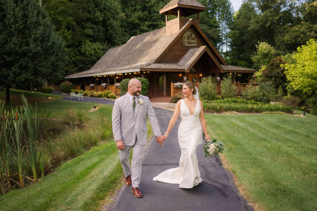 A joyful couple walks hand in hand down the picturesque pathway in front of the rustic Chapel in the Glen, surrounded by lush greenery and serene pond views, capturing the charm of a romantic Gatlinburg elopement.