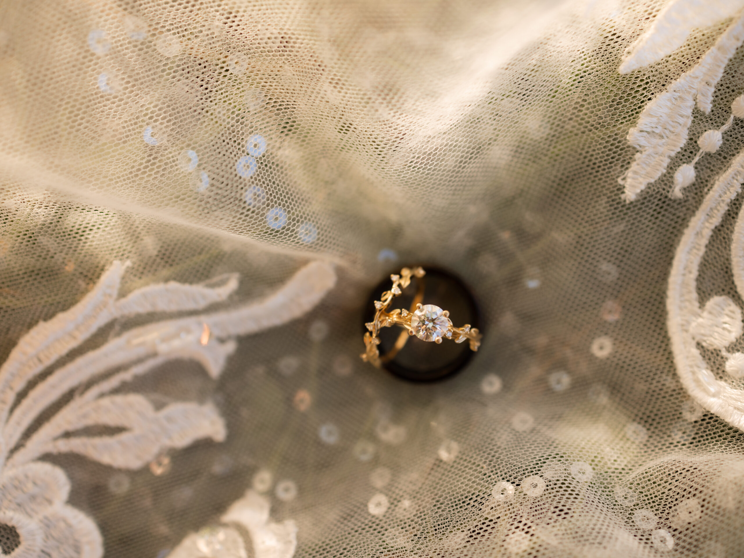 A stunning close-up of non-traditional wedding rings set against an intricate lace veil backdrop, softly illuminated by natural light. The bride’s ring features a delicate gold band with a nature-inspired design and a dazzling round diamond, while the groom’s simple dark band adds a striking contrast. Perfect for a Gatlinburg elopement.