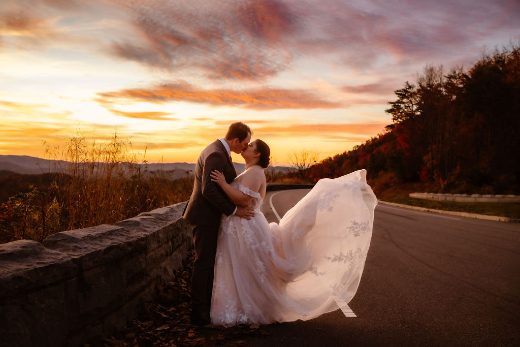 The couple embraces on a scenic mountain road as the bride’s flowing lace gown catches the breeze, framed by a fiery sunset and the autumnal beauty of Gatlinburg, captured by a Gatlinburg photographer during their elopement.