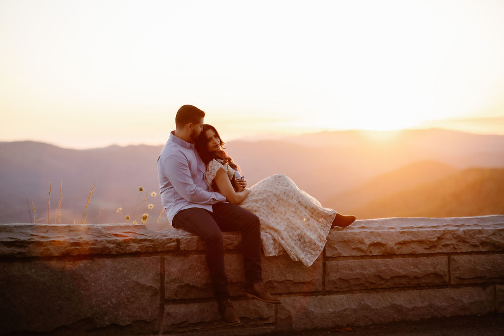 A couple sits on a stone wall overlooking the Great Smoky Mountains at sunset during their honeymoon. The man, dressed in a light blue shirt and dark pants, embraces the woman, who wears a flowing floral dress captured by Gatlinburg photographer