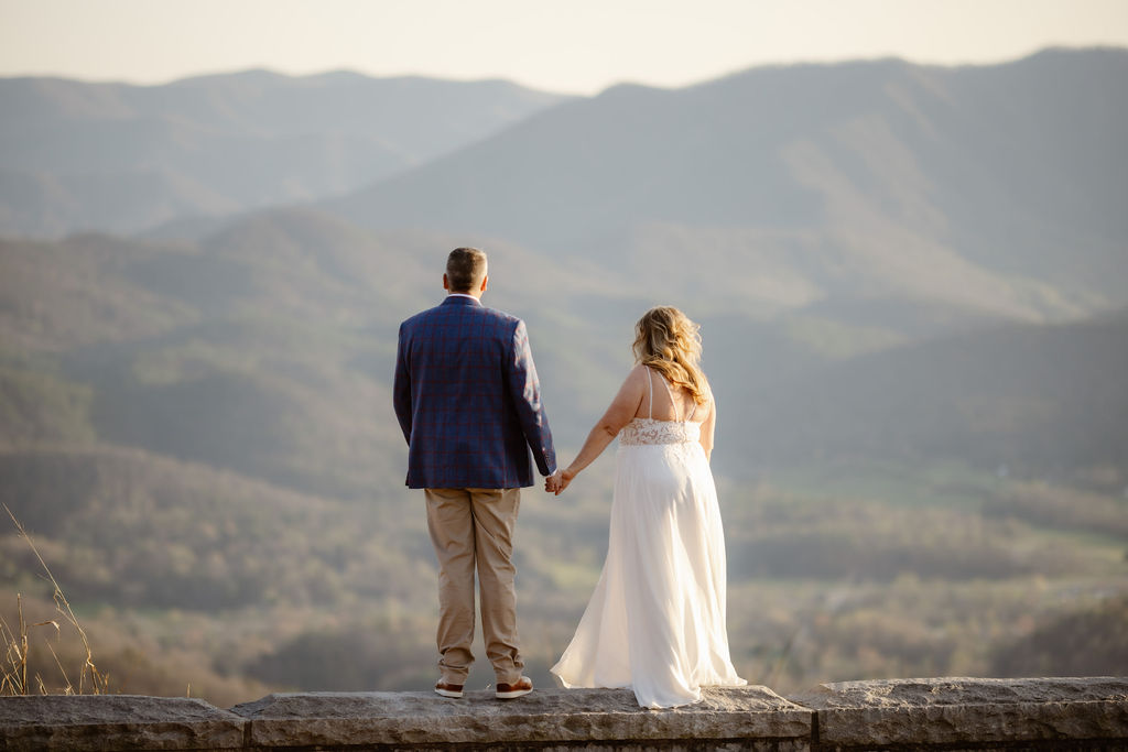 Bride and groom hold hands while looking out over the expansive Smoky Mountains from a stone ledge, representing the breathtaking views and peaceful moments offered by Gatlinburg Tennessee chapels for couples who elope to Gatlinburg.