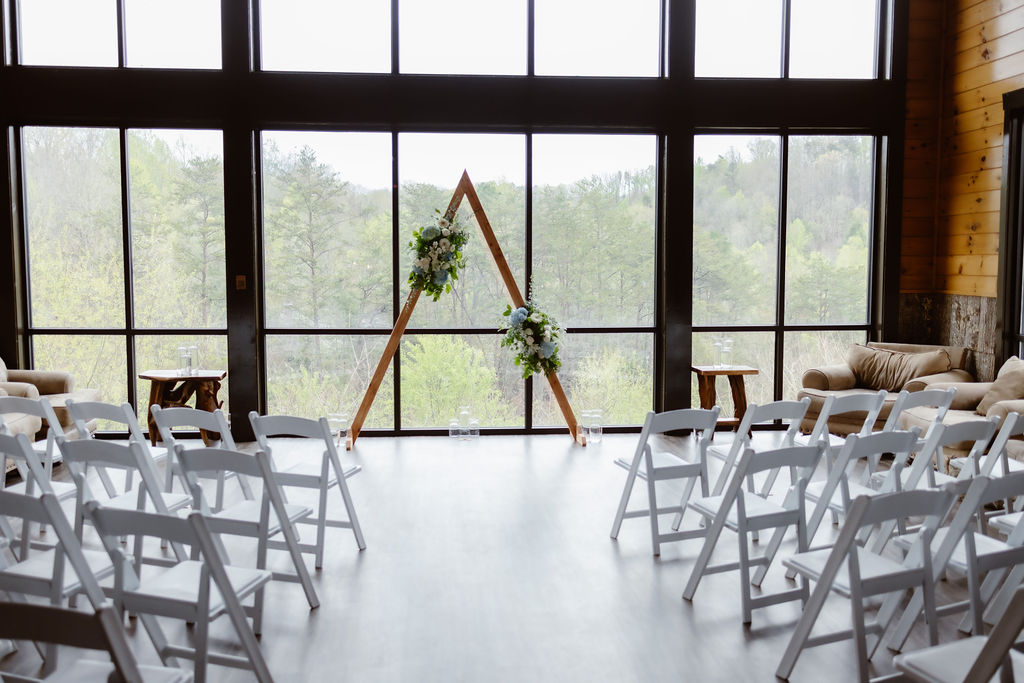 Indoor ceremony setup at Wild Briar Lodge featuring a wooden triangle arch adorned with greenery and florals, white chairs, and panoramic forest views—an inviting scene for couples eloping to Gatlinburg.