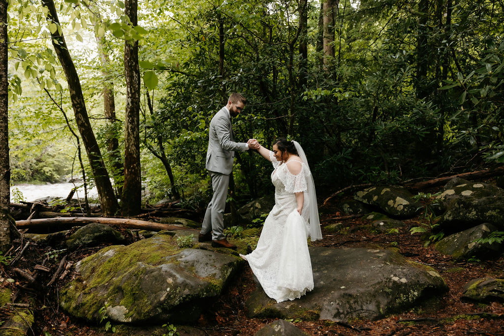 Bride and groom in wedding attire standing on moss-covered rocks in a forest near a river in Elkmont, Tennessee, with the groom helping the bride balance as she lifts her dress, captured by a Gatlinburg photographer.