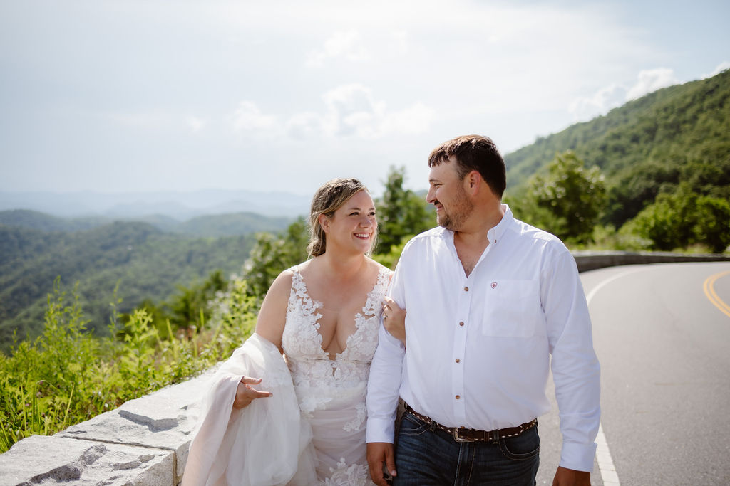 Bride and groom walking arm in arm along a scenic mountain road near Gatlinburg, Tennessee, with lush green hills and blue sky in the background, capturing a joyful moment from their decision to elope to Gatlinburg.