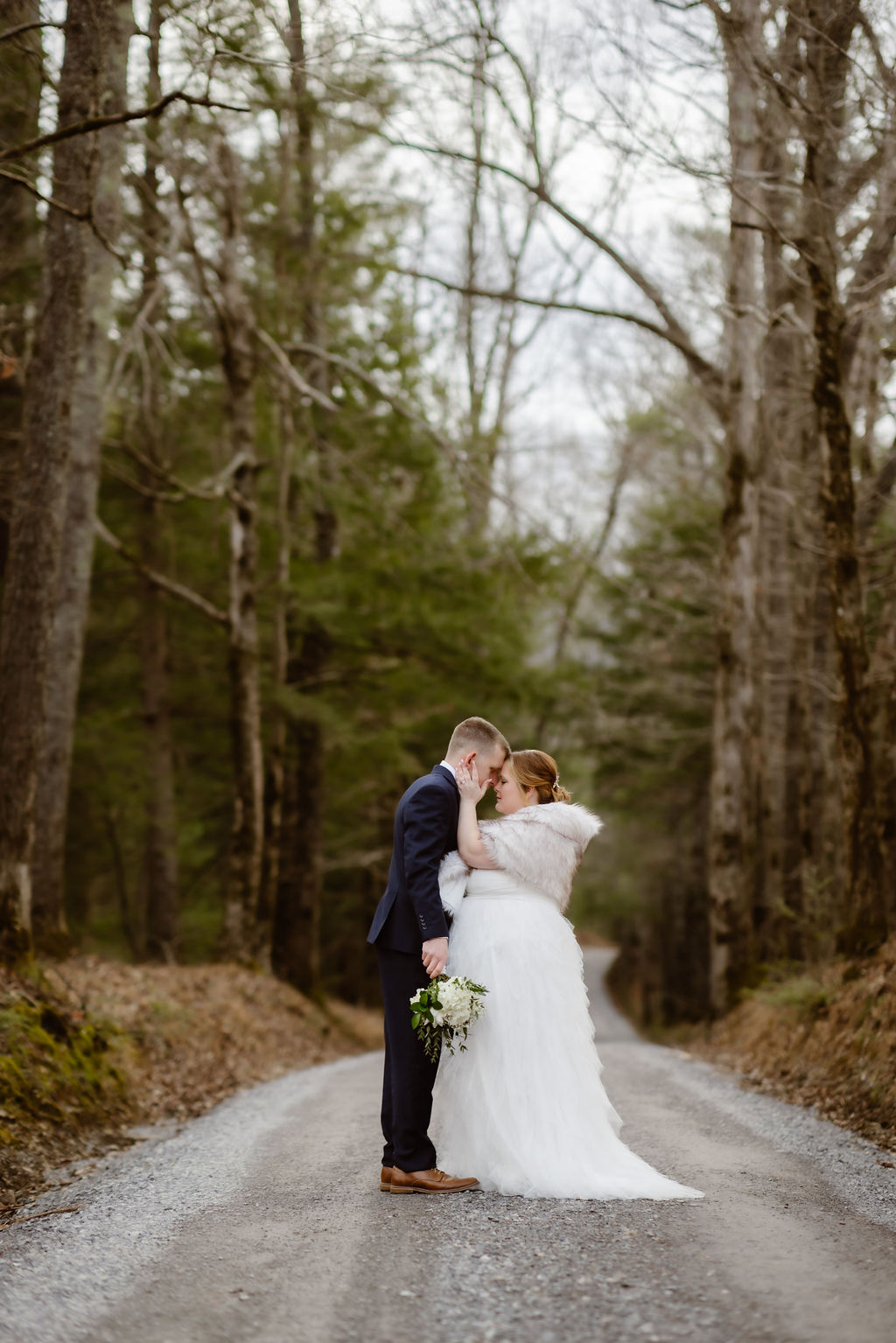 Bride and groom sharing an intimate moment on a secluded forest road in Cades Cove, her winter gown and bouquet highlighting the charm of couples who choose to elope to Gatlinburg in the winter, photographed by a Gatlinburg photographer.