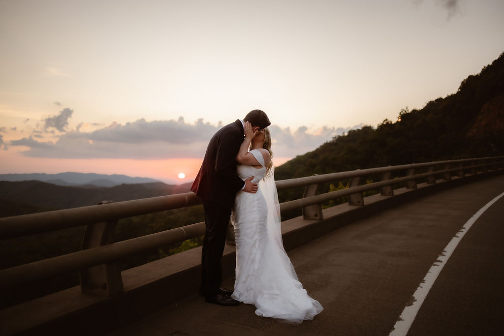 Couple eloping to Gatlinburg at Mountain Mist Farm wedding venue sharing a romantic kiss on a winding mountain road at sunset, with soft light casting a warm glow over the Smoky Mountain horizon.