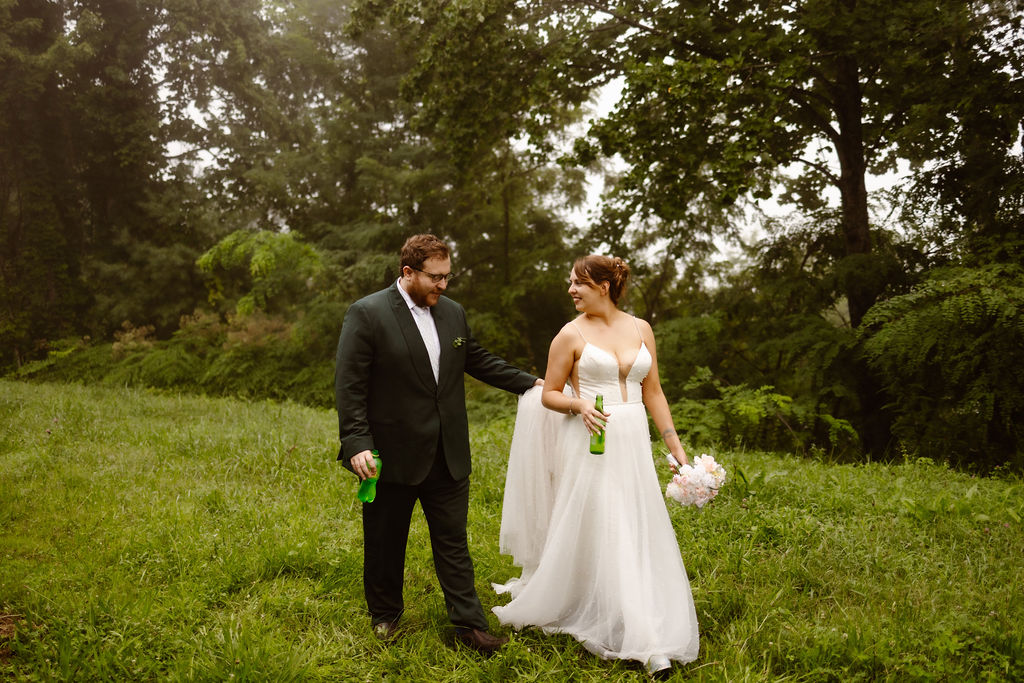 Bride and groom walking hand in hand through a lush green forest clearing, each holding a drink, after their Great Smoky Mountain wedding ceremony, captured by Gatlinburg photographer.