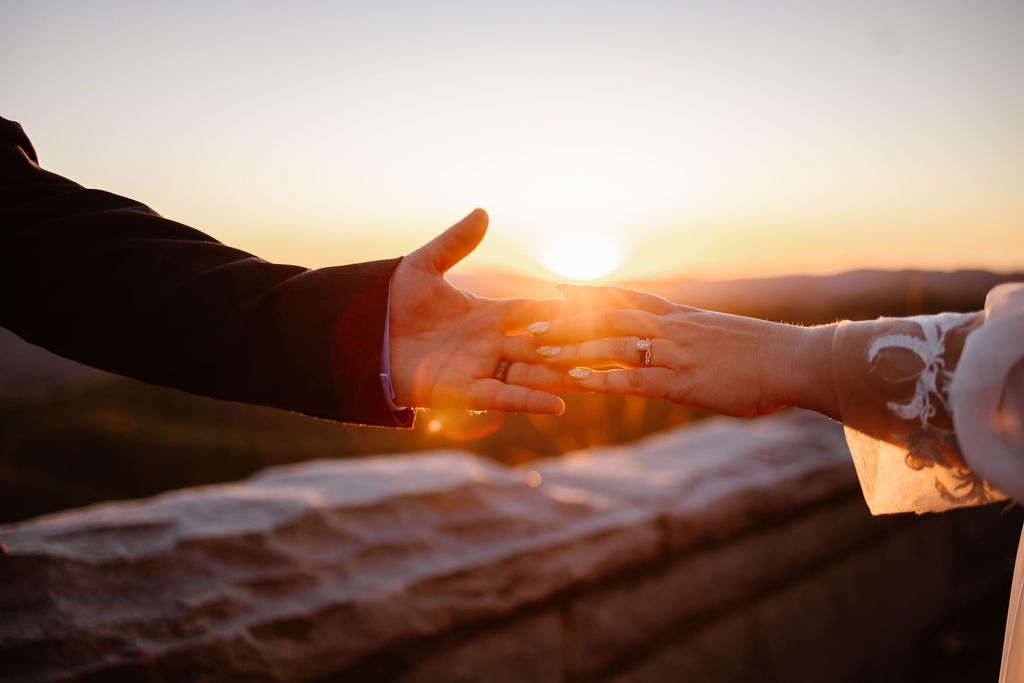 Close-up of the couple’s hands reaching for each other in front of a stone ledge, with a wedding ring visible and the sun setting in the distance during a gatlinburg elopement