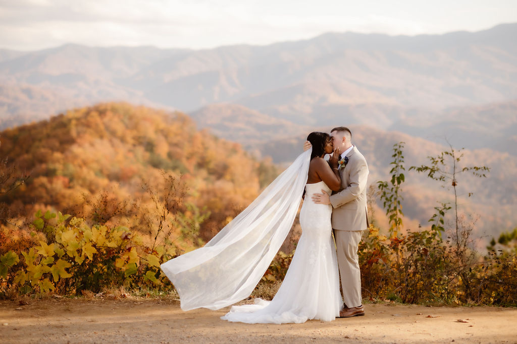 Bride and groom share a kiss on a mountain overlook with fall foliage and flowing veil, highlighting the scenic appeal of a Gatlinburg wedding venue and the intimate experience of eloping to Gatlinburg.
