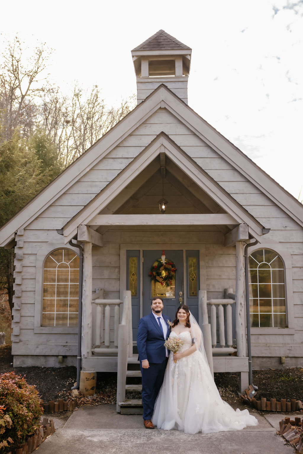 Bride and groom standing in front of a small rustic chapel with a holiday wreath on the door in Gatlinburg, Tennessee, showcasing a cozy and charming setting typical of Gatlinburg Tennessee chapels.