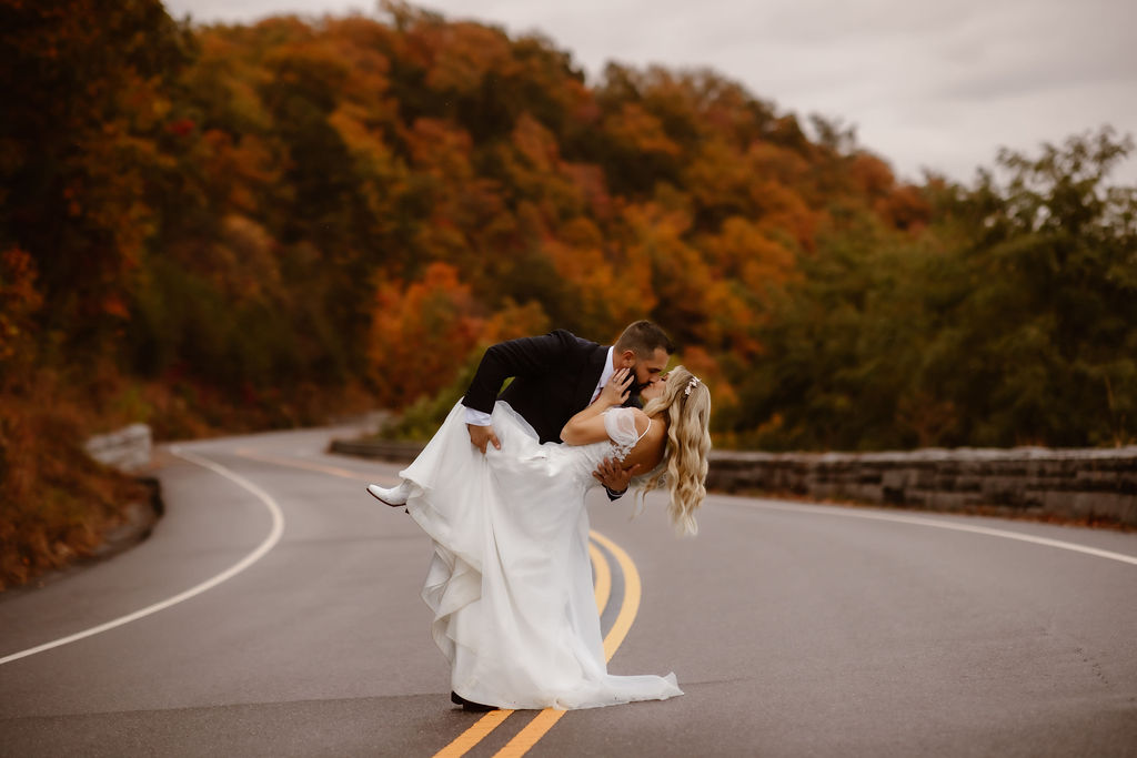 Groom dips and kisses bride in the middle of a winding mountain road lined with vibrant fall foliage, captured by Smoky Mountain photographers during an adventurous wedding portrait session.