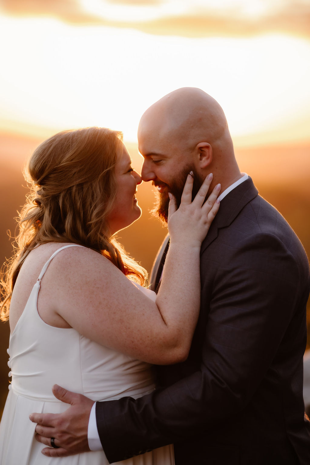 Intimate golden hour portrait of the newlyweds embracing at a scenic smoky mountain overlook, their faces softly lit by the setting sun with glowing Appalachian views behind them.