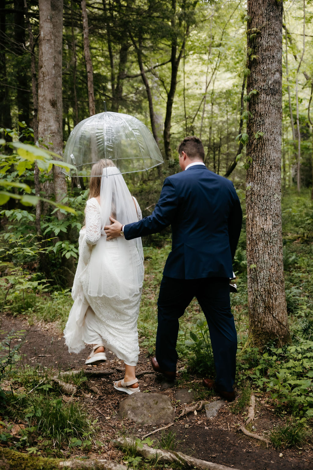 Bride and groom walk together beneath a clear umbrella along a wooded trail in Elkmont, surrounded by mossy trees and soft forest light. This intimate moment captures the natural charm of eloping in Gatlinburg, even with a touch of mountain rain.