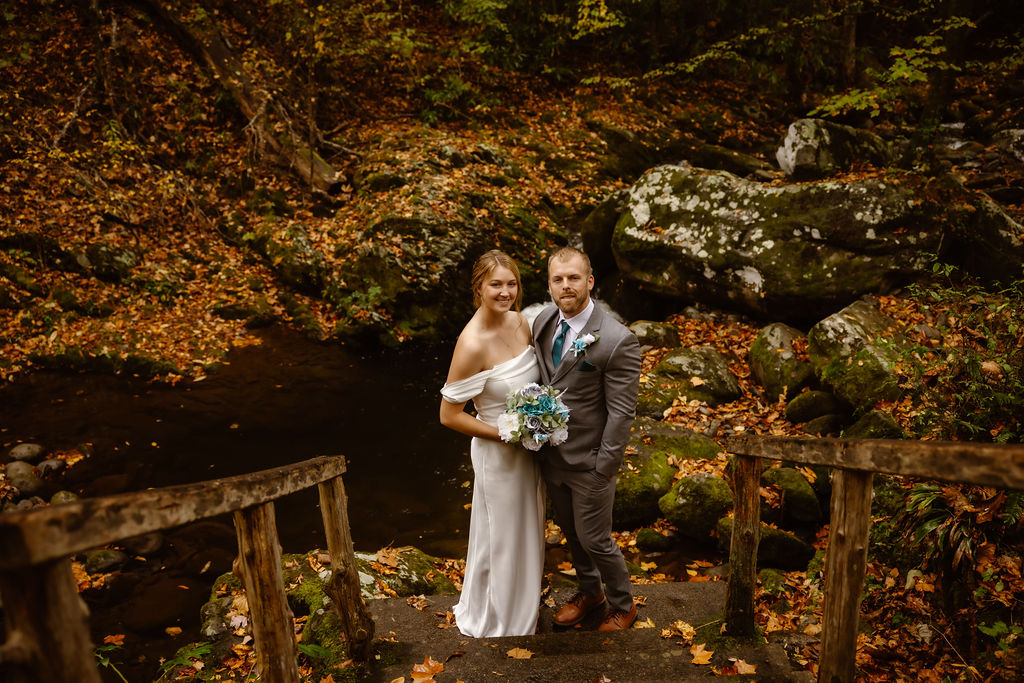 Bride and groom standing on a rustic wooden bridge over a stream during a water fall elopement at Ely's Mill, captured in a scenic Gatlinburg elopement setting in the Smoky Mountains with autumn foliage and moss-covered rocks