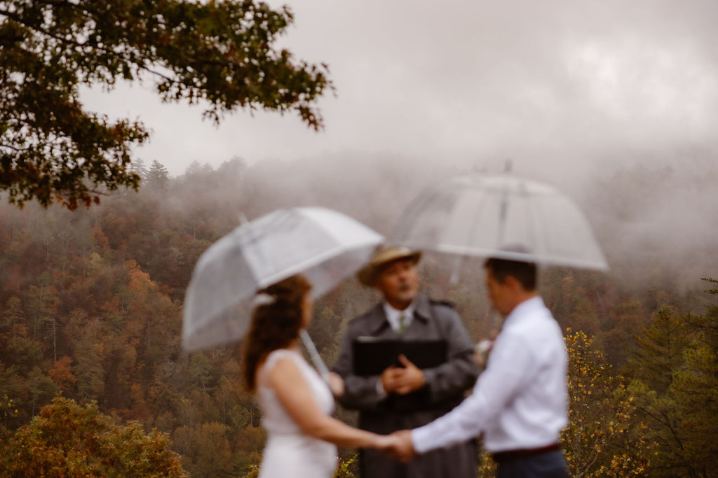Rainy wedding day ceremony during a Gatlinburg elopement with a couple exchanging vows beneath clear umbrellas, surrounded by mist-covered mountain views and autumn foliage, capturing romantic outdoor wedding planning inspiration in a moody natural setting.