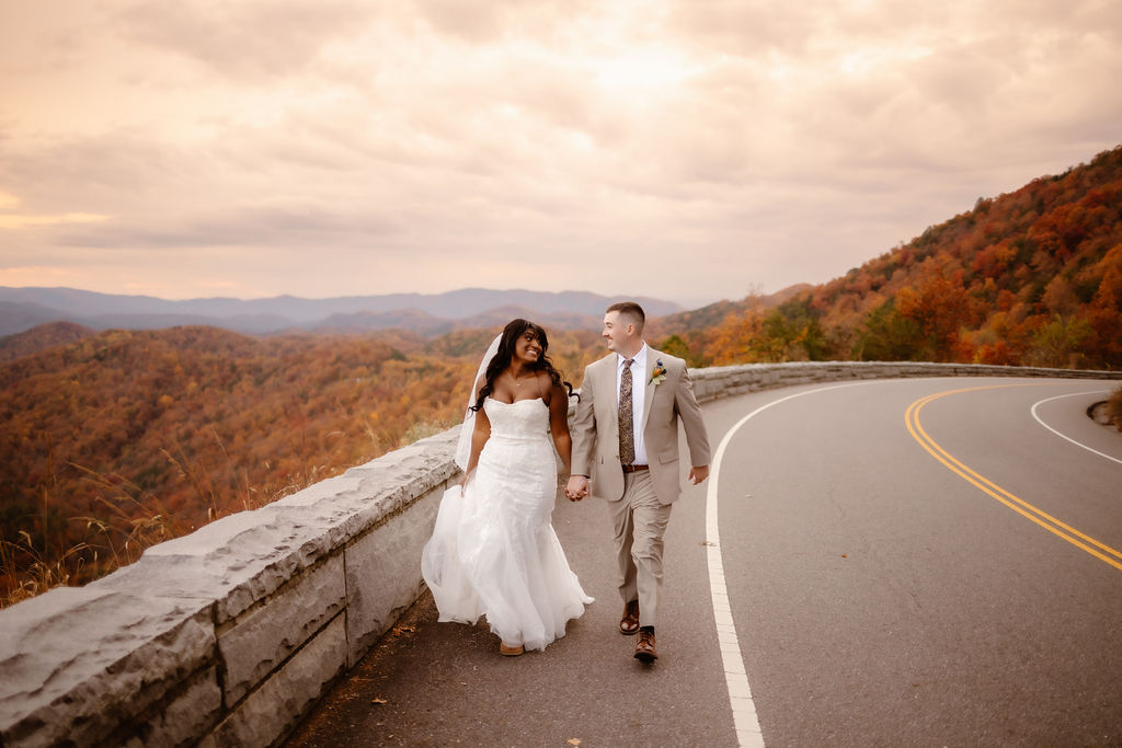 Bride and groom walking hand in hand along a mountain overlook road during a Smoky Mountain elopement with colorful fall foliage in the background.