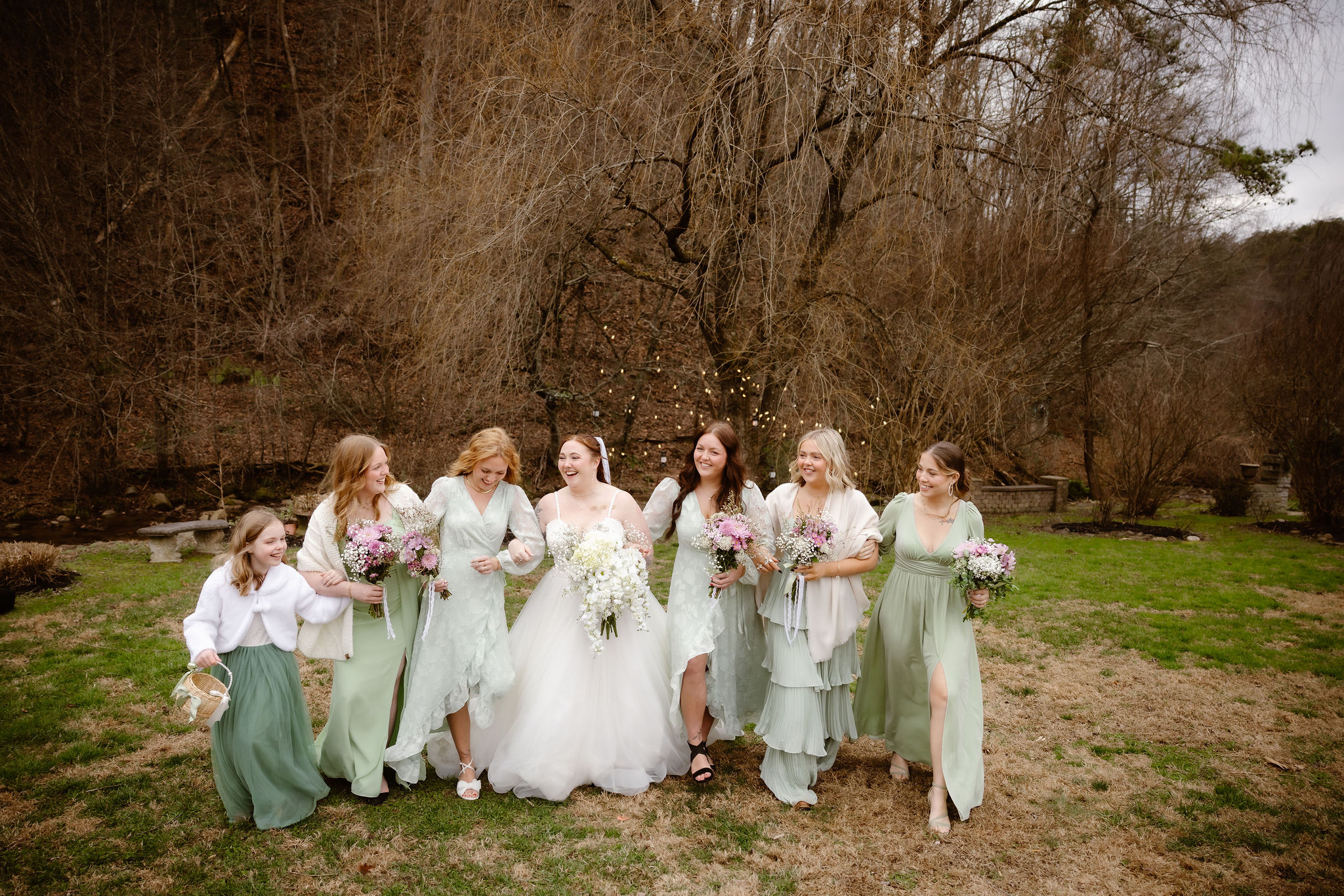 Bride in a white wedding gown walking arm-in-arm with six bridesmaids in sage green dresses and a flower girl carrying a basket during a **Gatlinburg elopement**, holding pastel floral bouquets on a grassy lawn with towering leafless trees and a wooded mountain backdrop.
