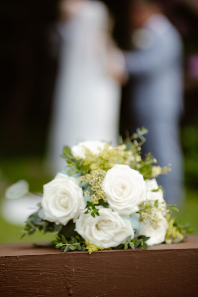 White rose bridal bouquet resting on a wooden railing with the newlyweds softly blurred in the background at Honeysuckle Hills.
