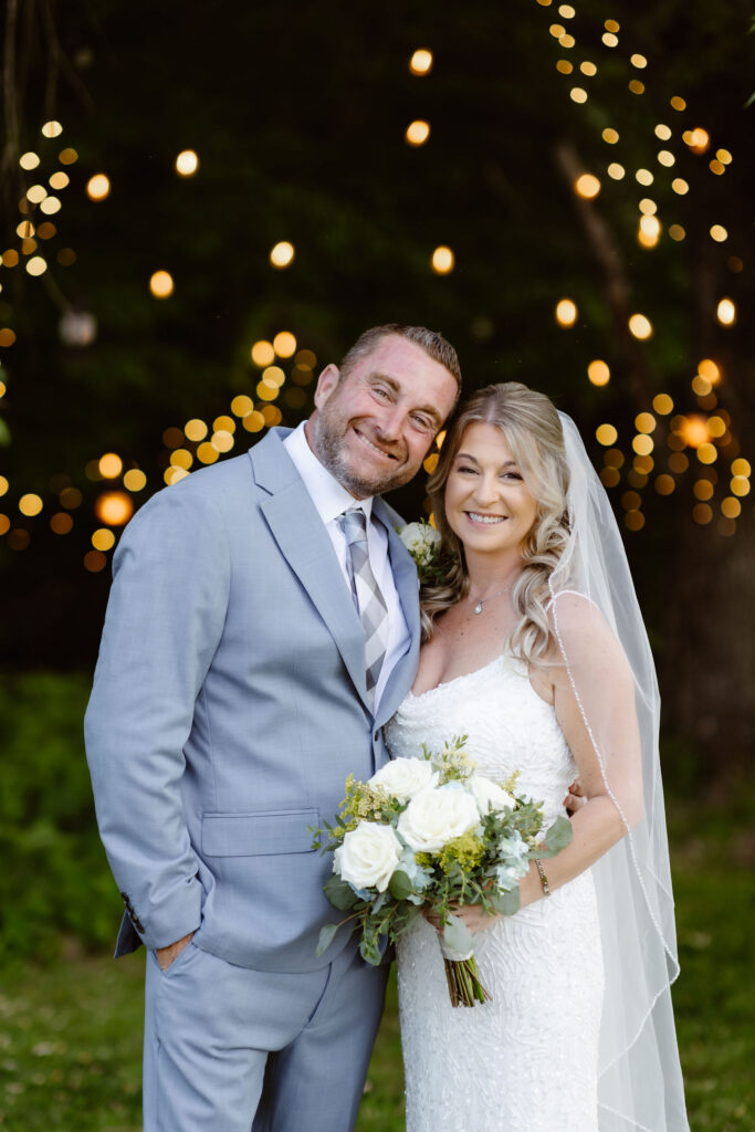 Bride and groom smiling together beneath glowing string lights at Honeysuckle Hills after their wedding ceremony.