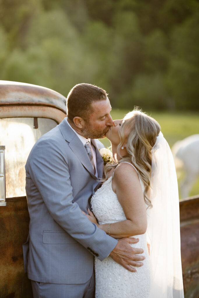 Bride and groom sharing a kiss beside a vintage truck at Honeysuckle Hills during golden hour, with horses grazing in the background.