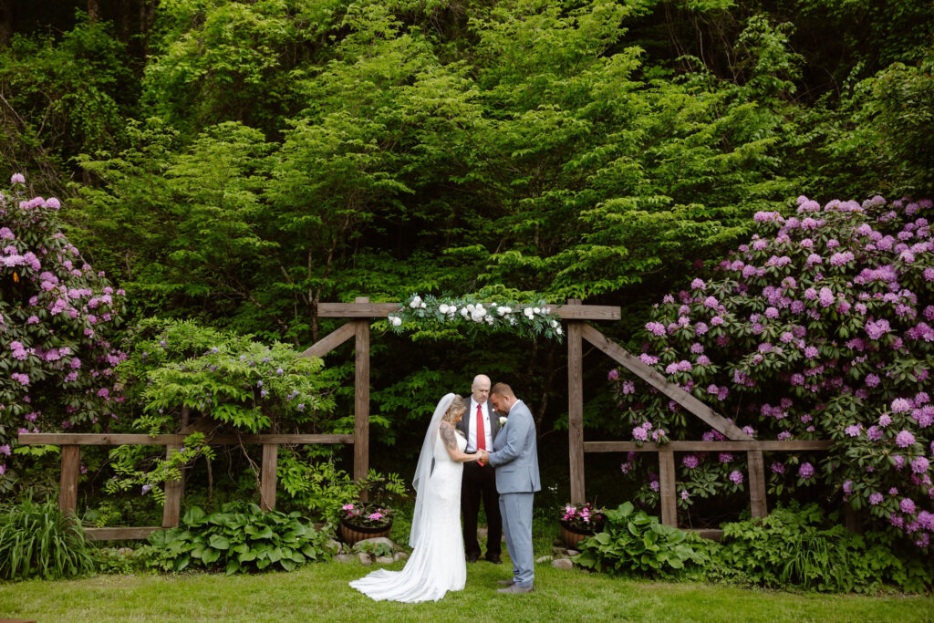 Bride and groom exchanging vows beneath a wooden arbor surrounded by rhododendrons at Honeysuckle Hills, a peaceful Gatlinburg wedding venue.