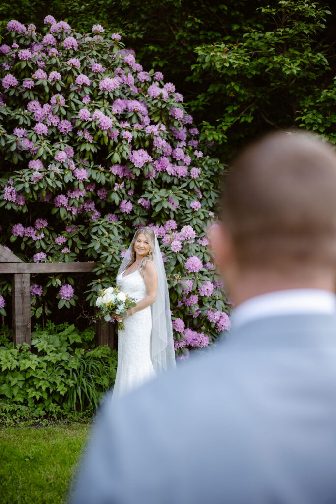 Bride holding a bouquet in front of blooming rhododendrons as the groom watches during their first look at Honeysuckle Hills.