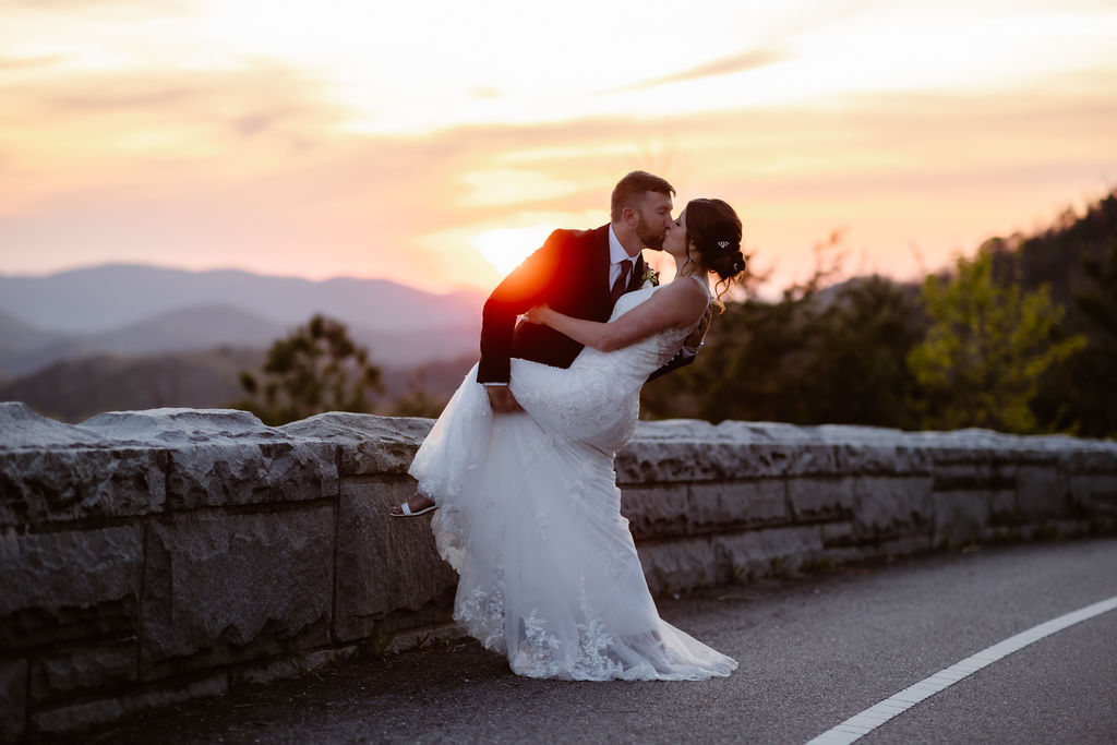 Groom dipping the bride for a kiss during a sunset wedding at Foothills Parkway, captured by a Gatlinburg photographer with the Smoky Mountains glowing in the background