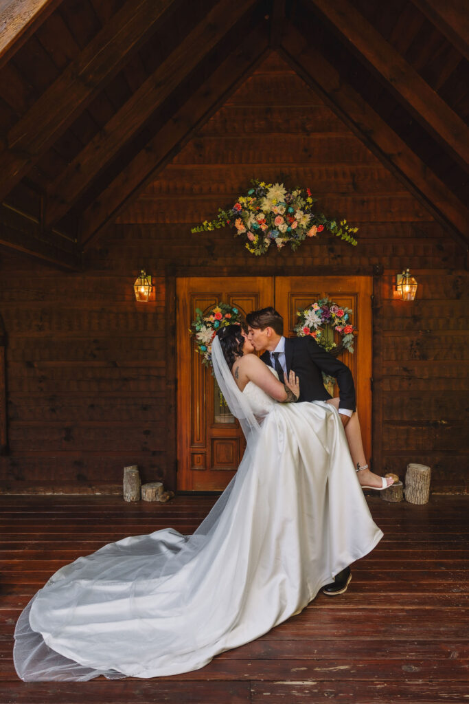 Newlyweds share a playful dip and kiss beneath colorful floral wreaths outside the Little Log Chapel in Gatlinburg after their intimate ceremony.