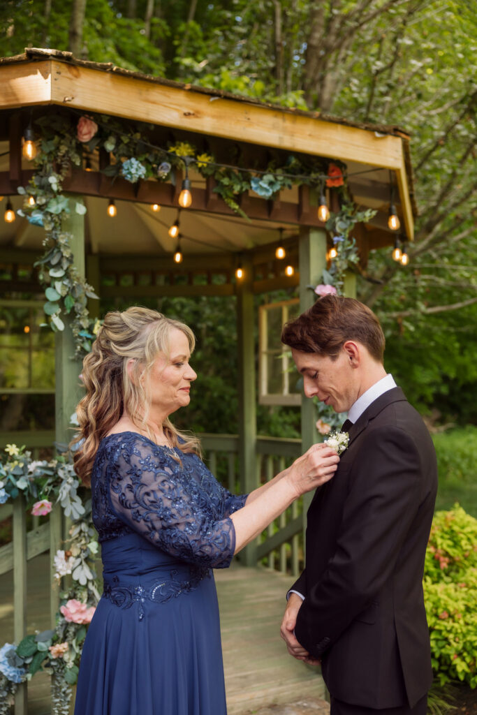 Woman in a blue lace dress pins a white boutonniere on the groom beside a flower-covered gazebo during a Gatlinburg elopement.
