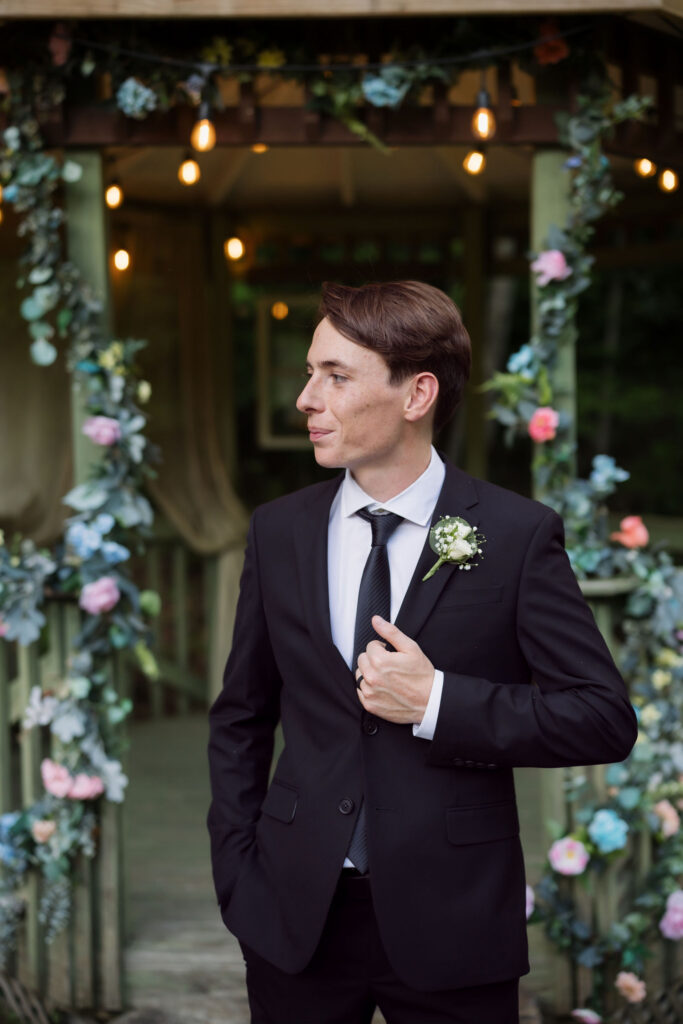 Groom in a black suit poses beneath string lights and floral garlands during a Gatlinburg elopement at the Little Log Chapel in Gatlinburg.